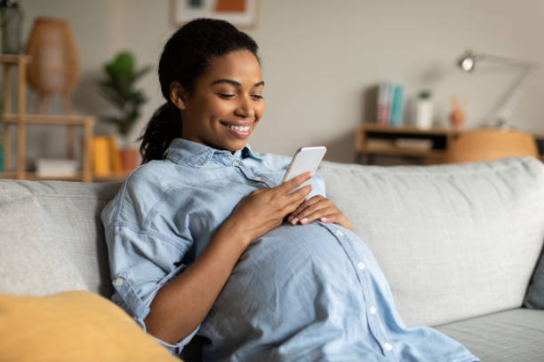 Dekorationsbild: Gravid kvinna sitter i soffa ler och tittar på sin mobiltelefon